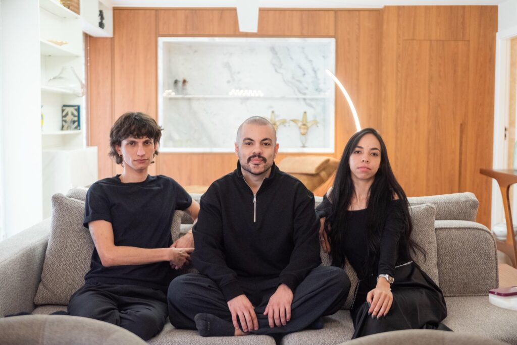 Três integrantes da equipe Guima sentados em um sofá, em uma sala com painel em madeira